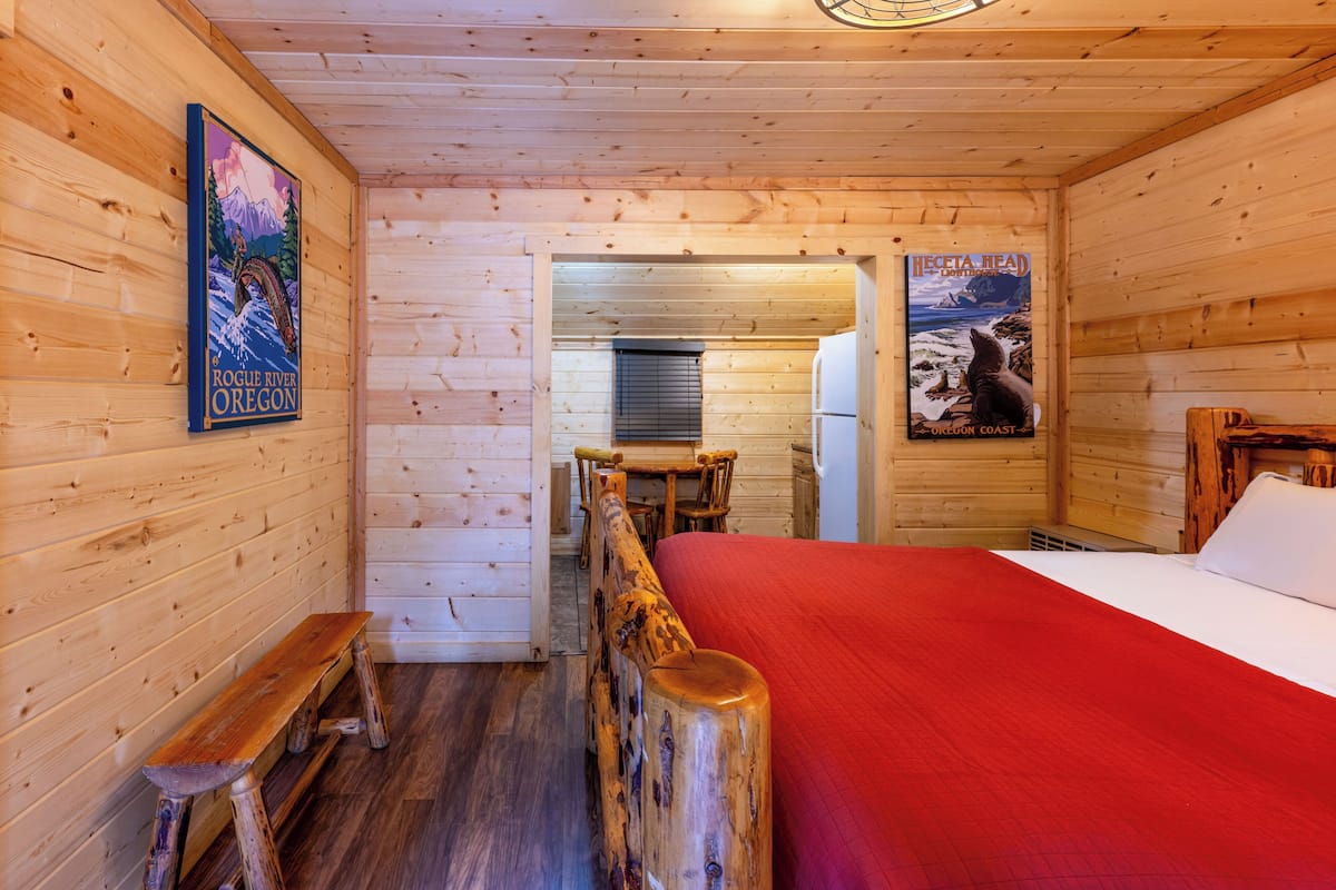 Interior de cabana de madeira com cama, banco, paredes de madeira e cartazes.