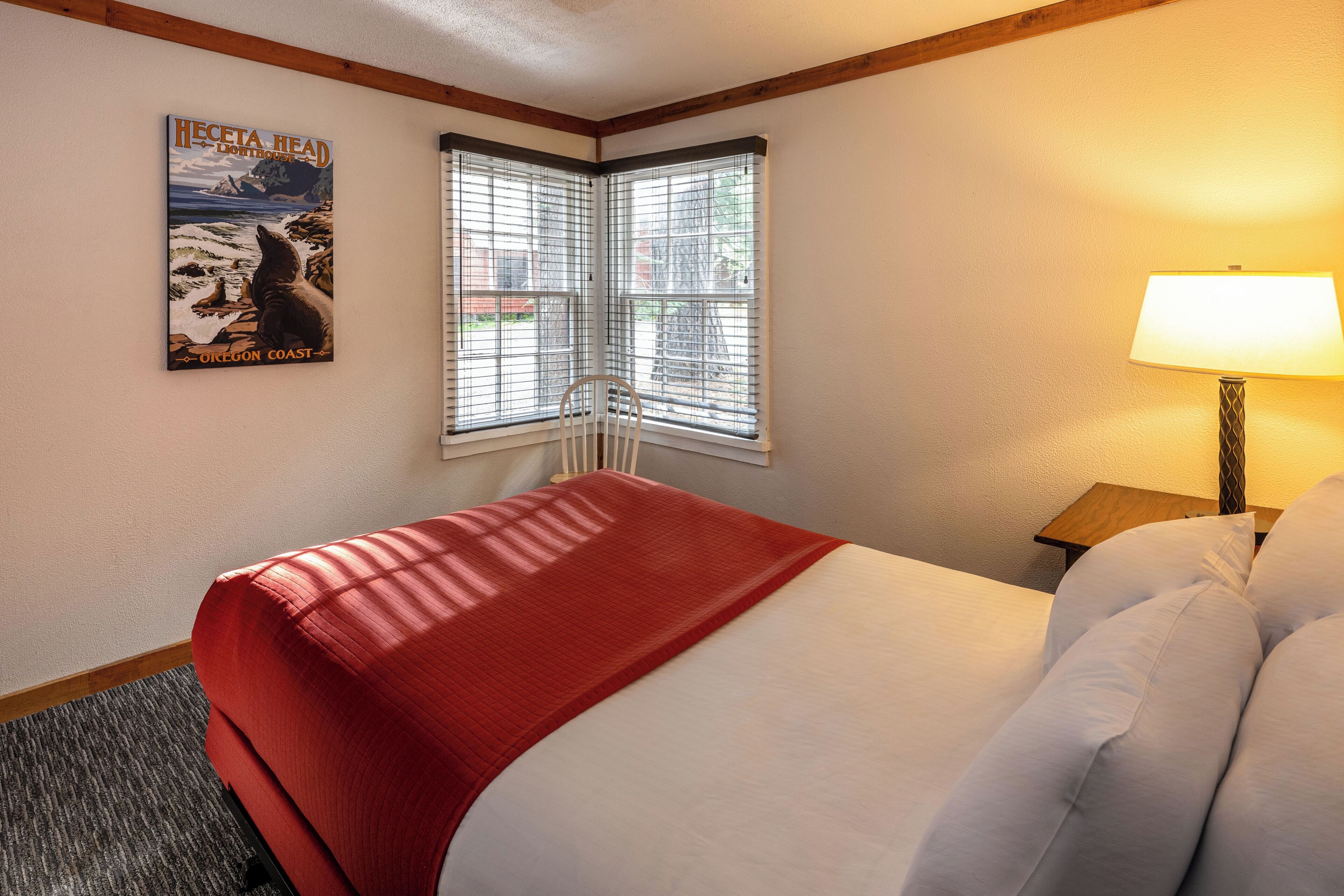 Ein Hotelzimmer mit Bett, einem Fenster mit Jalousien, einer Lampe und einem Poster an der Wand.