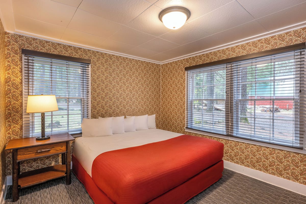 Quarto de hotel com uma cama grande, duas janelas com cortinas, uma mesa de cabeceira com abajur e papel de parede estampado.