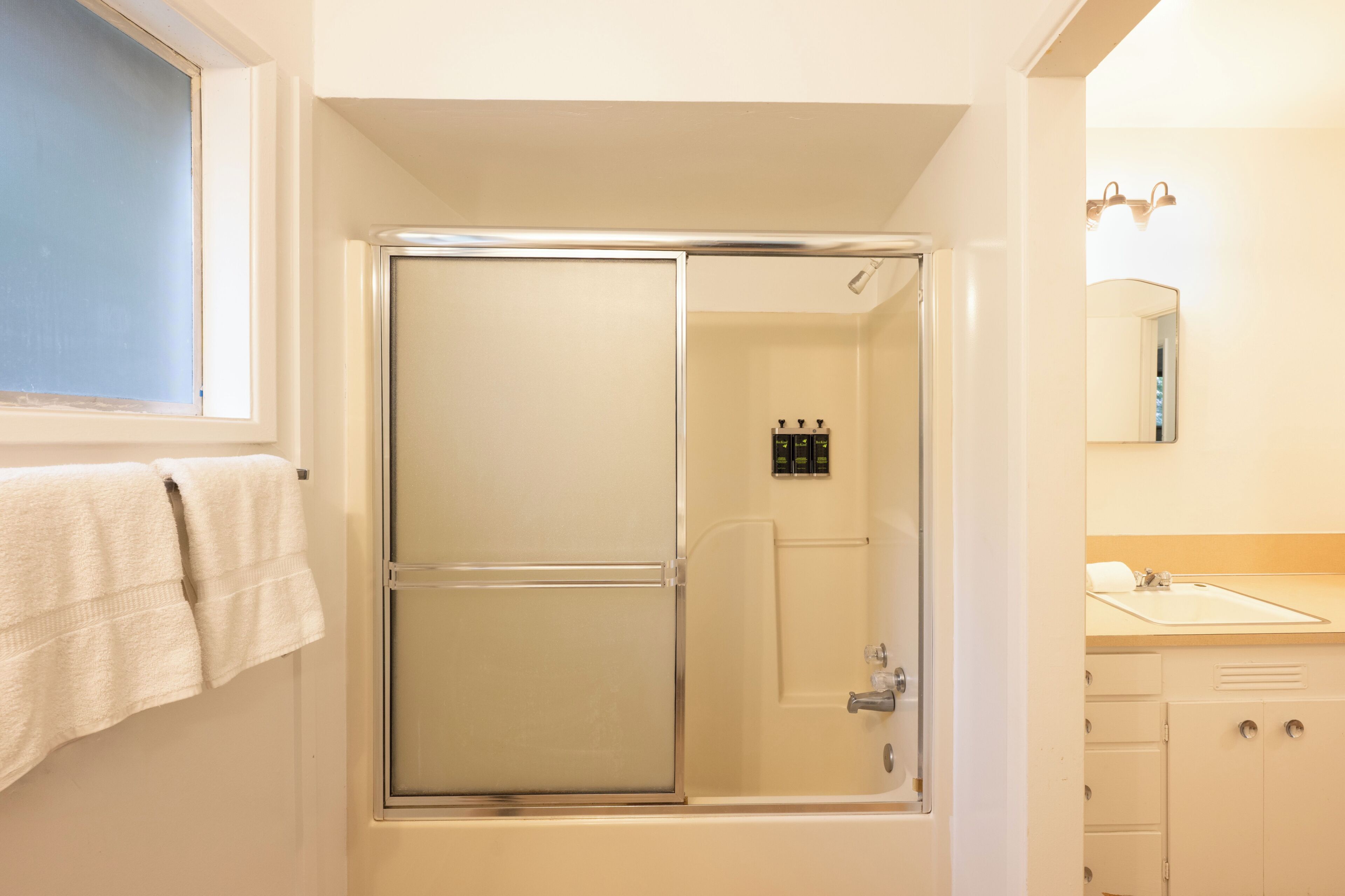 A bathroom with a glass-enclosed shower, a window with a towel, and a sink with a mirror.