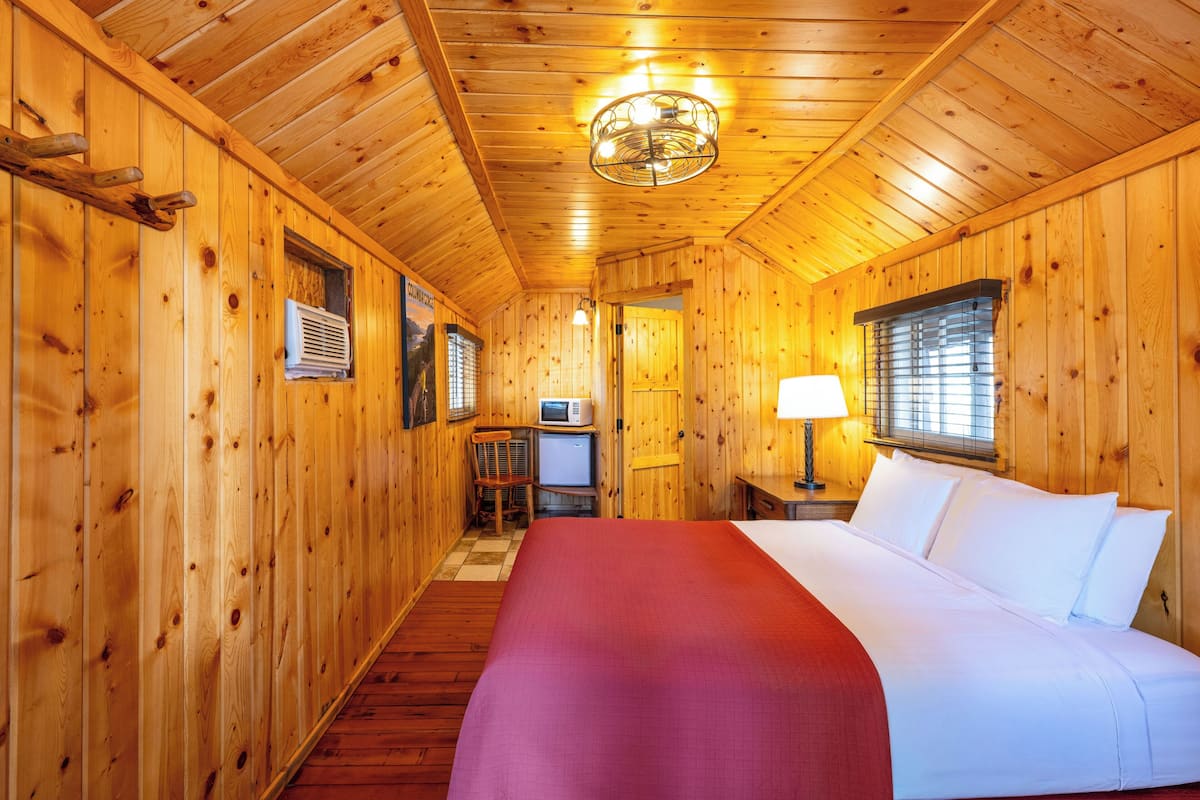 Quarto de cabana de madeira com cama, escrivaninha, cadeira, televisão pequena e janela com persianas.