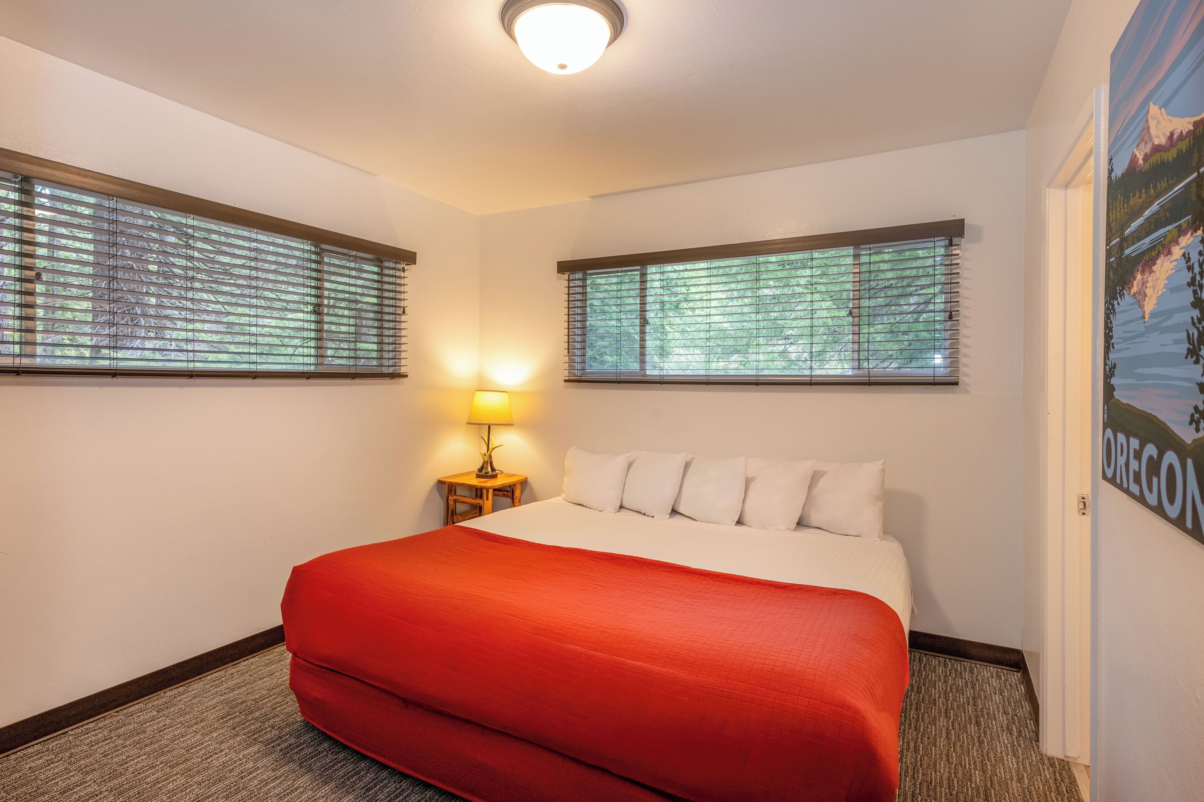 A hotel room with a bed, two windows with blinds, a nightstand with a lamp, and a poster on the wall.