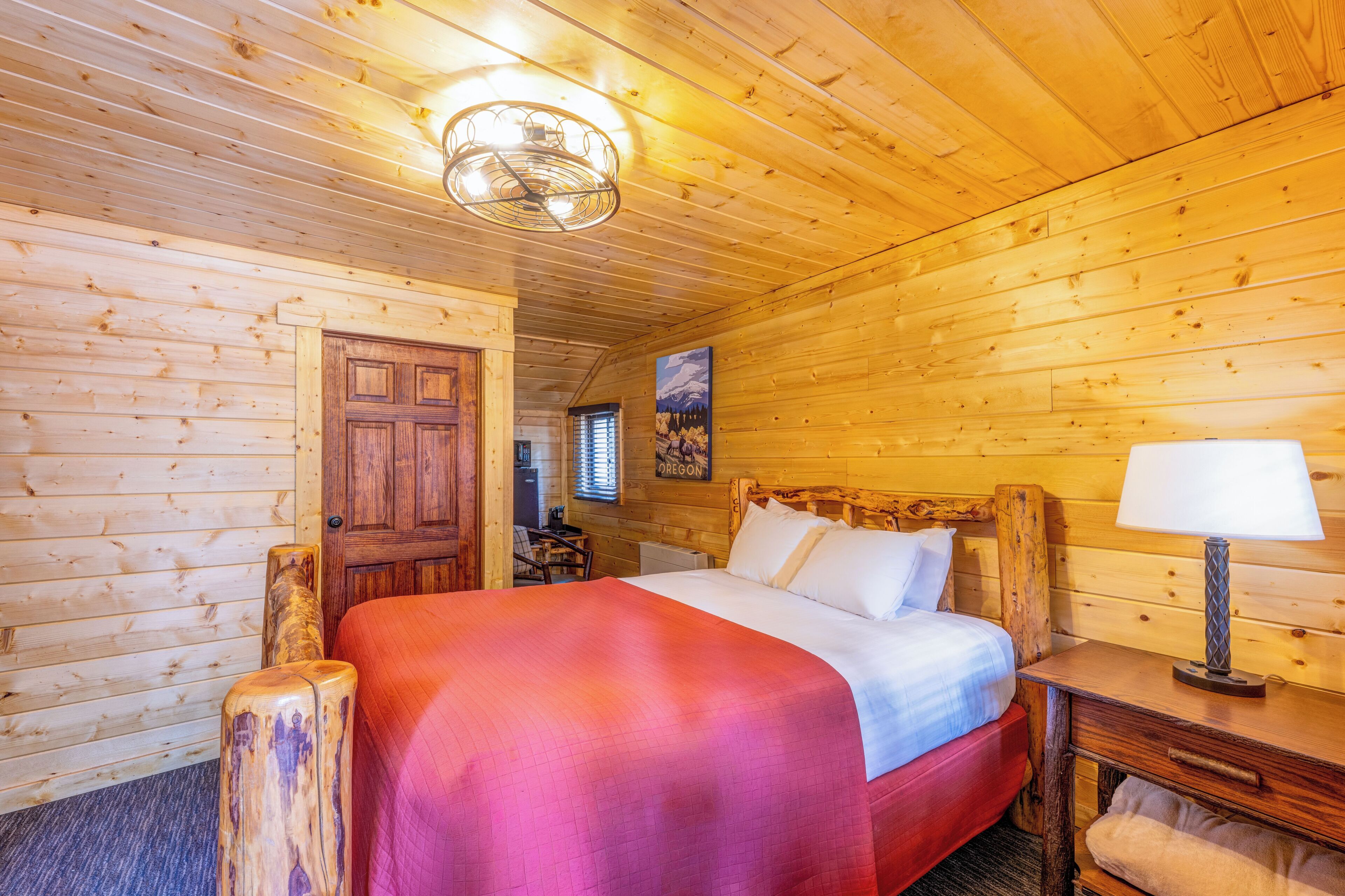 Una habitación tipo cabaña de madera con una cama, cabecera de madera, mesita de noche, lámpara y una puerta.