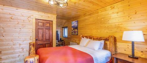 Una habitación tipo cabaña de madera con una cama, cabecera de madera, mesita de noche, lámpara y una puerta.