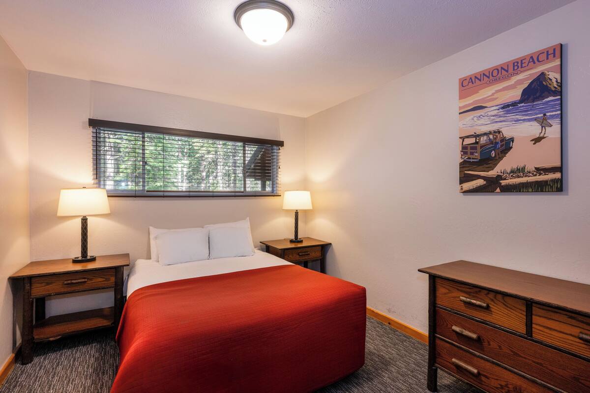 Quarto de hotel com uma cama, duas mesas de cabeceira com luminárias, uma cômoda, uma janela com persianas e um pôster na parede.