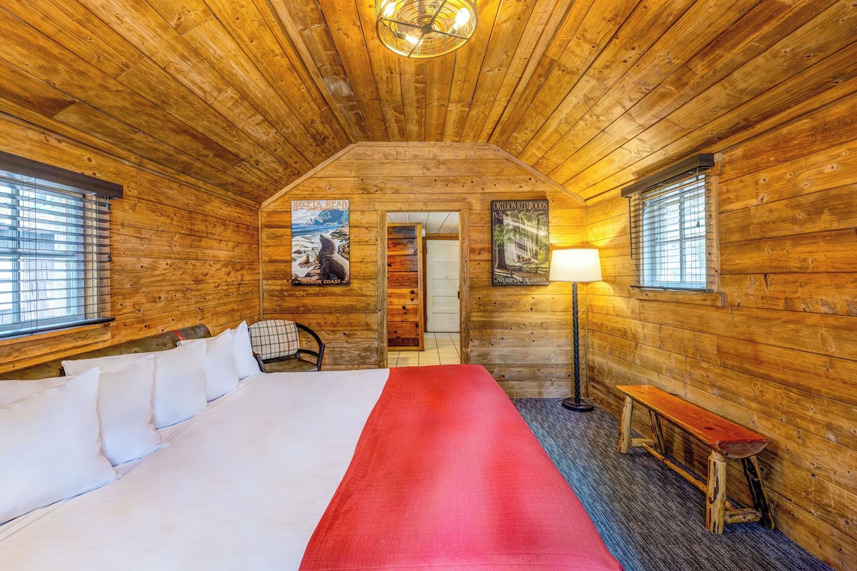 Quarto de cabana rústica com uma cama grande, um banco, um abajur e dois cartazes nas paredes.