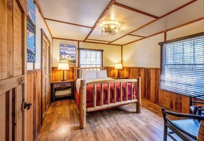 A wooden room with a bed, two lamps, a window with blinds, and a door.