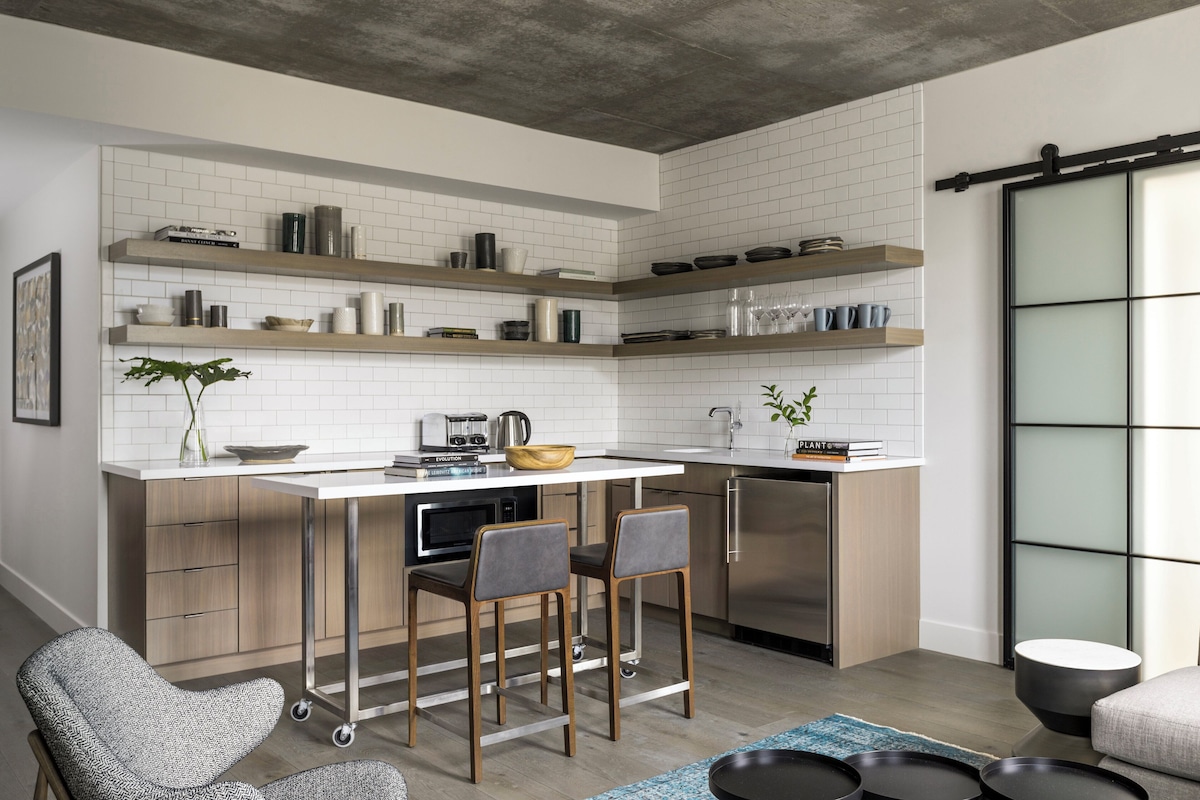 A modern kitchen with white subway tiles, wooden shelves, a central island with bar stools, and a built-in oven.