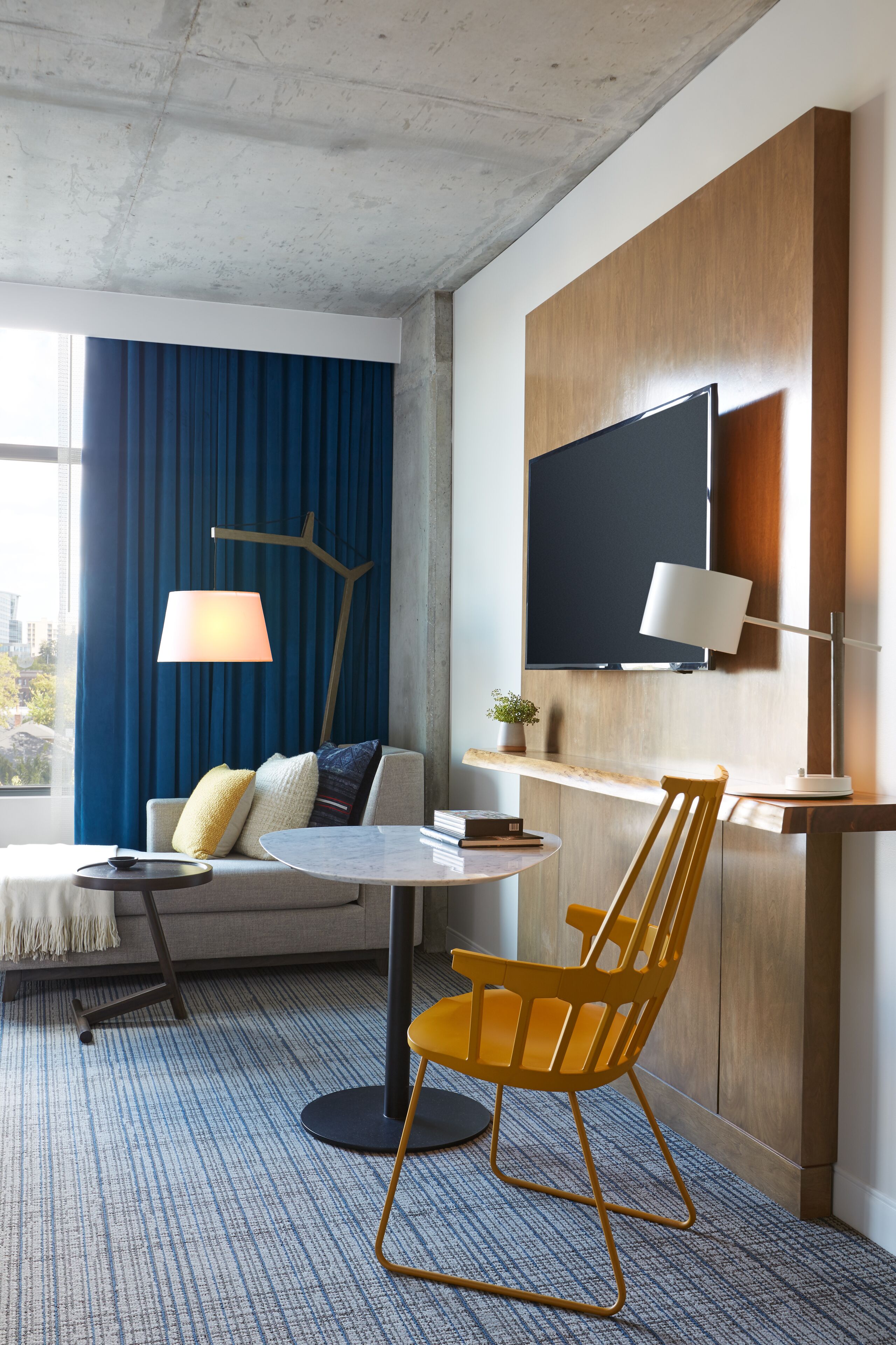 A modern hotel room with a sofa, a round table, a yellow chair, a TV mounted on the wall, and a large window with blue curtains.