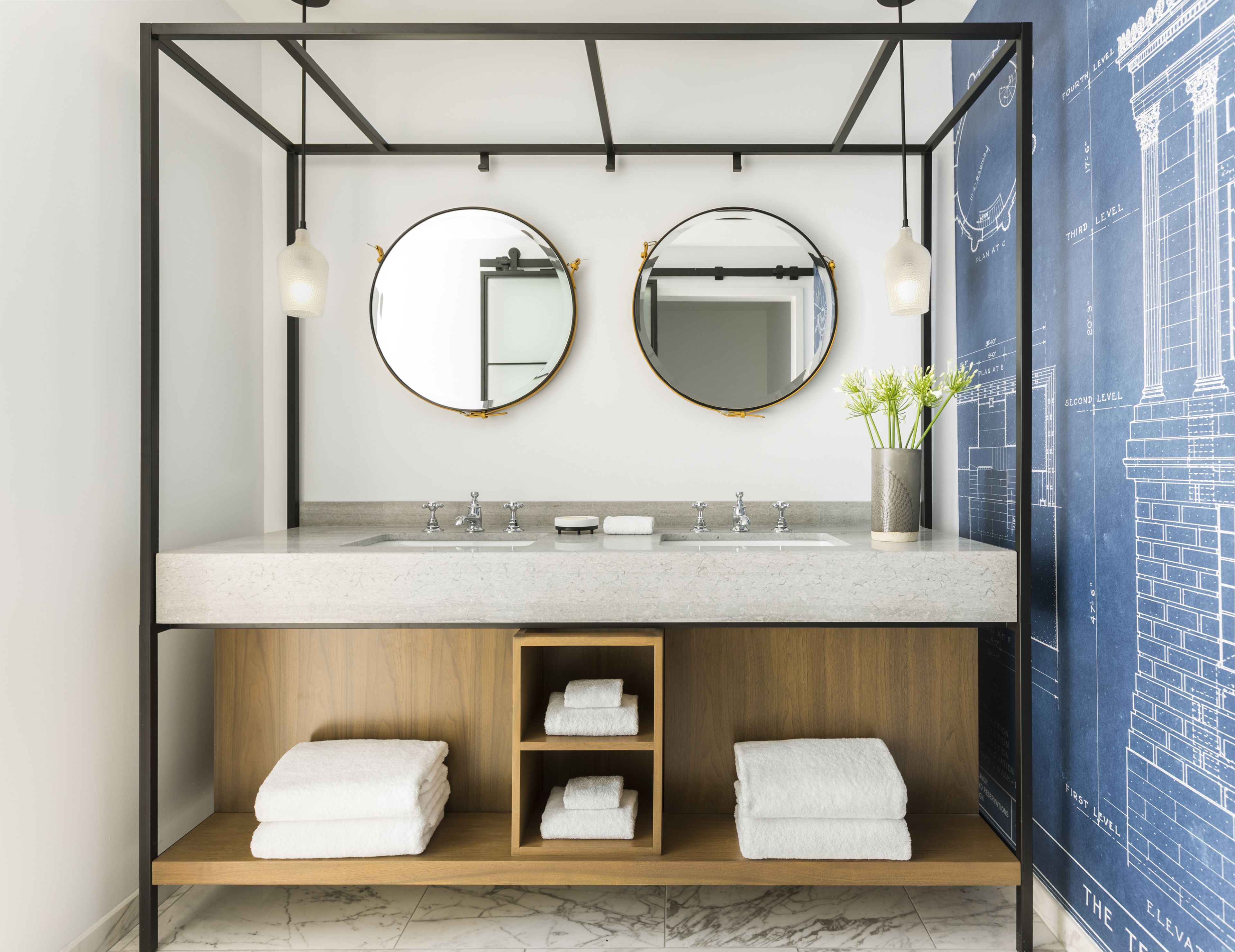 A modern bathroom with a double sink, two round mirrors, a vase with flowers, and a towel rack with folded towels.