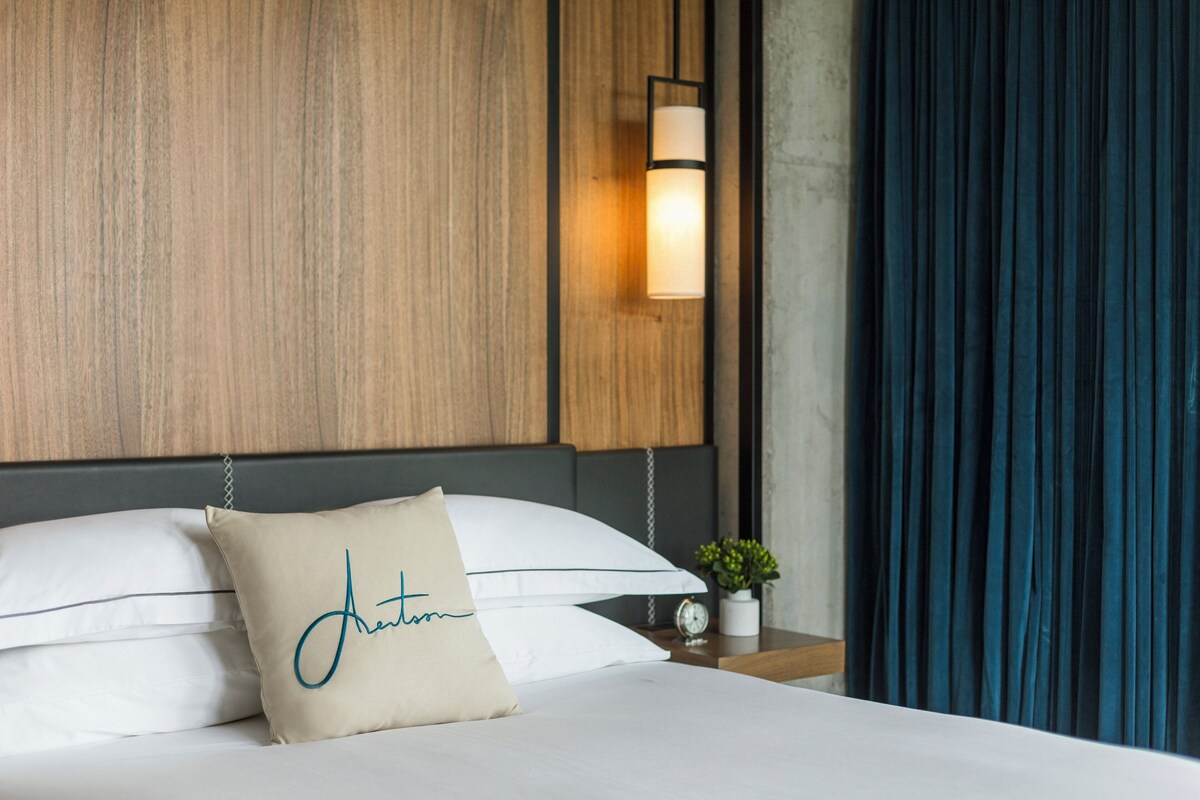 A neatly made bed with a pillow labeled 'Antonia,' a bedside table with a clock, and a wall-mounted lamp.