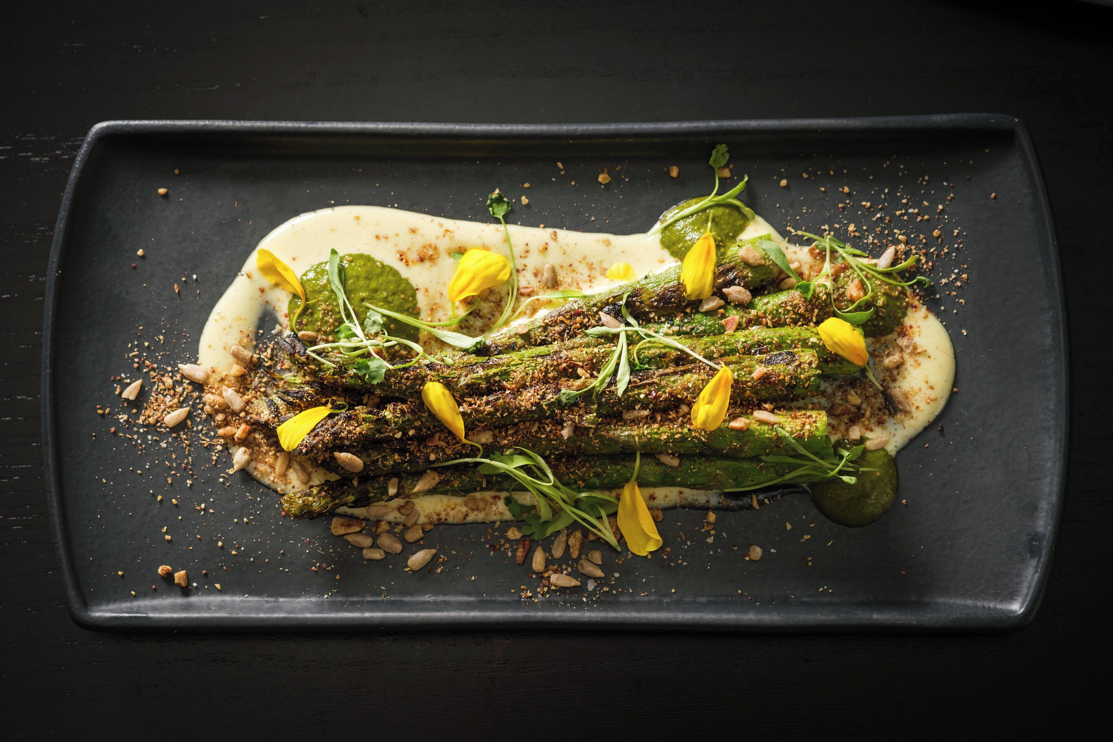A rectangular plate with a dish of grilled asparagus, garnished with herbs and lemon slices, on a dark background.