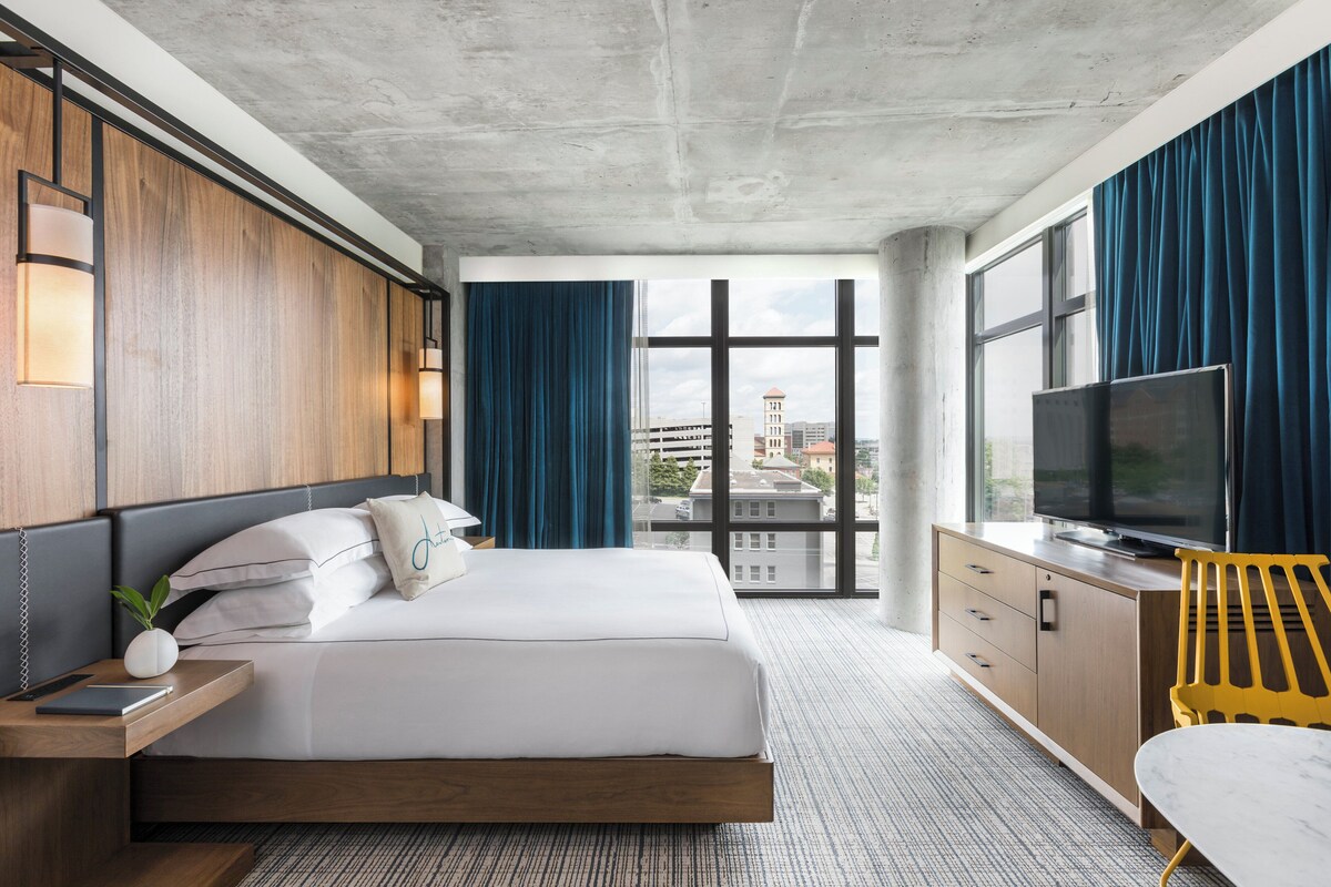 A modern hotel room with a large bed, a TV, a wooden nightstand, a desk, a chair, and a view of the city through large windows.