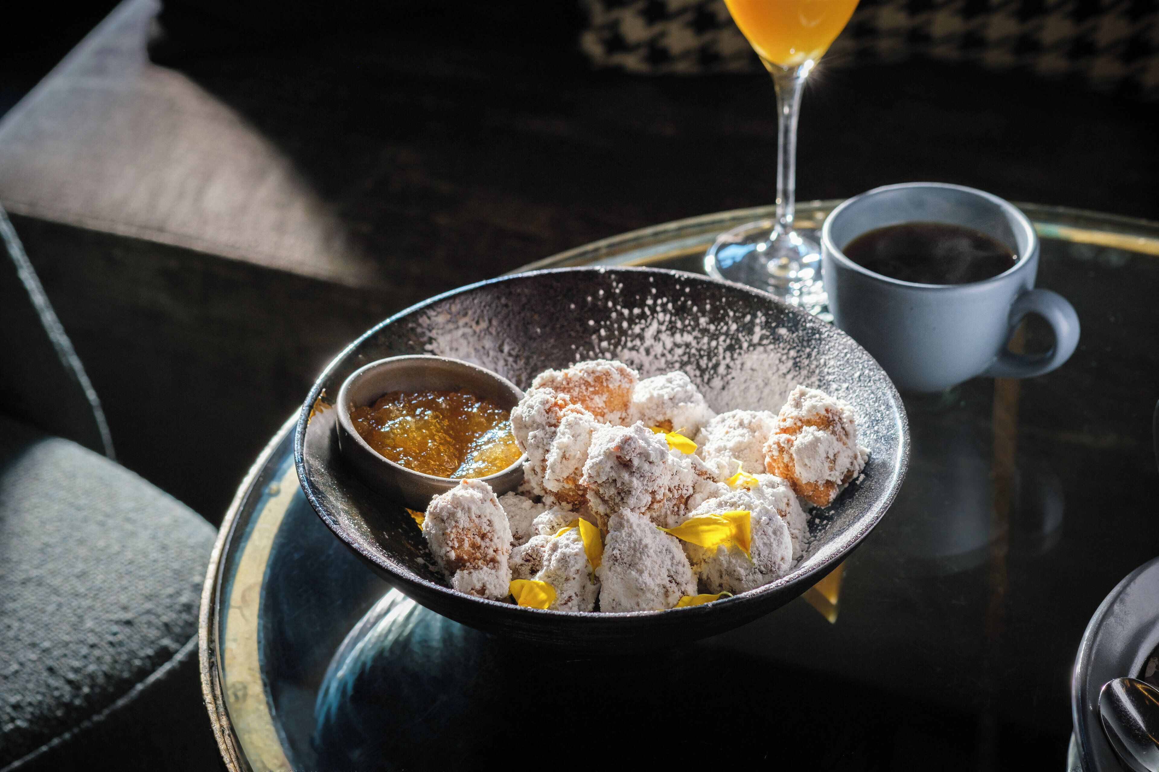 Un tazón de dulces cubiertos de azúcar glas, un recipiente pequeño con salsa, un vaso de jugo de naranja y una taza de café sobre una superficie reflectante.