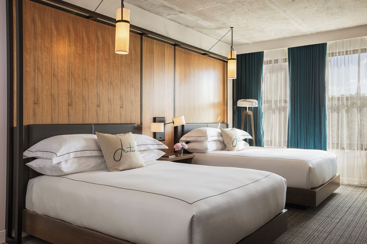 A hotel room with two beds, wooden paneling, a nightstand with a lamp, and a view of the city through sheer curtains.