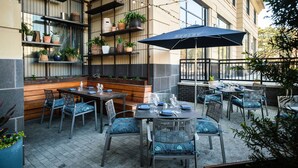 An outdoor dining area with tables and chairs, a large umbrella, and shelves with plants. - Kimpton Aertson Hotel by IHG (Nashville)