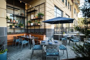 An outdoor dining area with tables and chairs, a large umbrella, and shelves with plants. - Kimpton Aertson Hotel by IHG (Nashville)