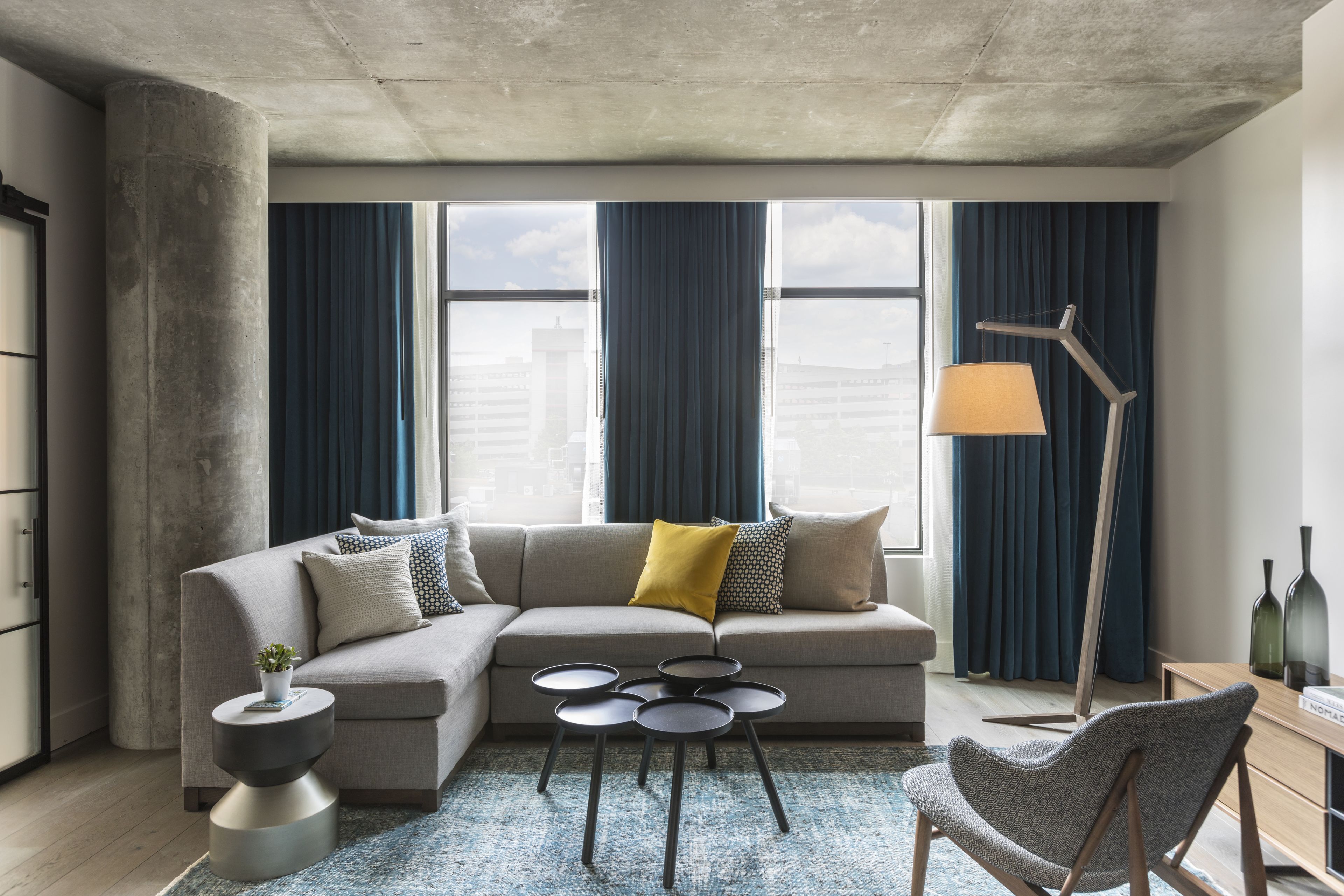 Ein modernes Wohnzimmer mit einem Ecksofa, einem Couchtisch, einem Beistelltisch, einer Stehlampe und Blick auf die Stadt durch große Fenster.