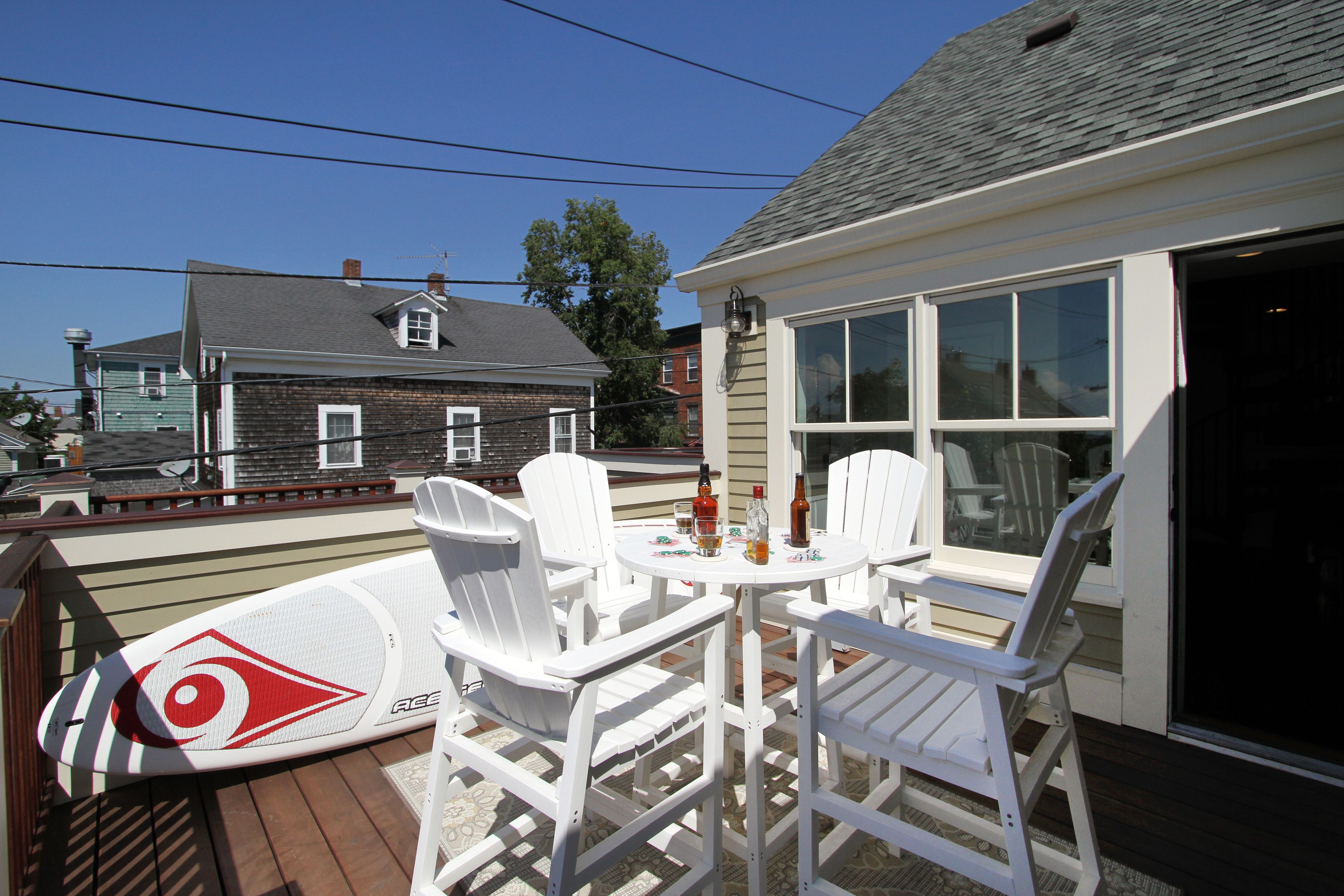 A deck with white chairs and a table, a surfboard, and a house in the background.