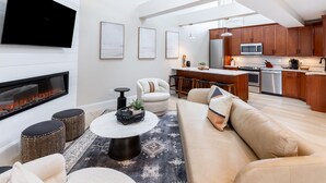 A modern living room with a fireplace, a sofa, a coffee table, and a kitchen area with wooden cabinets and stainless steel appliances. - The Newport Lofts – 543 Thames Street (Newport)