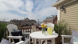 A deck with white furniture, a table with a pitcher of lemonade, and a view of houses in the background. - The Newport Lofts – 543 Thames Street (Newport)