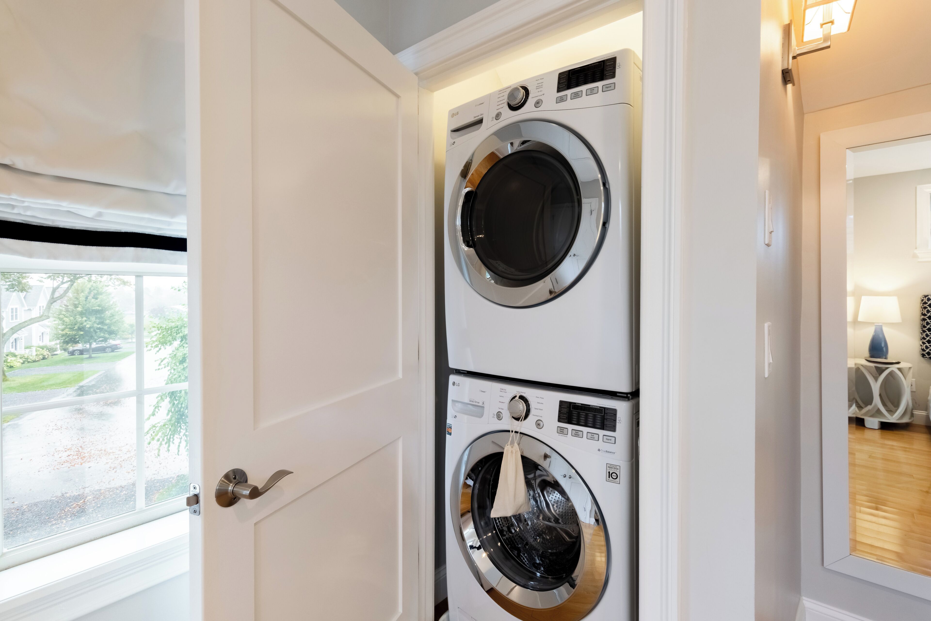 A laundry room with two stacked front-loading washing machines, a door with a silver handle, and a window with a view of a street and greenery.