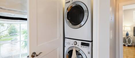 A laundry room with two stacked front-loading washing machines, a door with a silver handle, and a window with a view of a street and greenery.