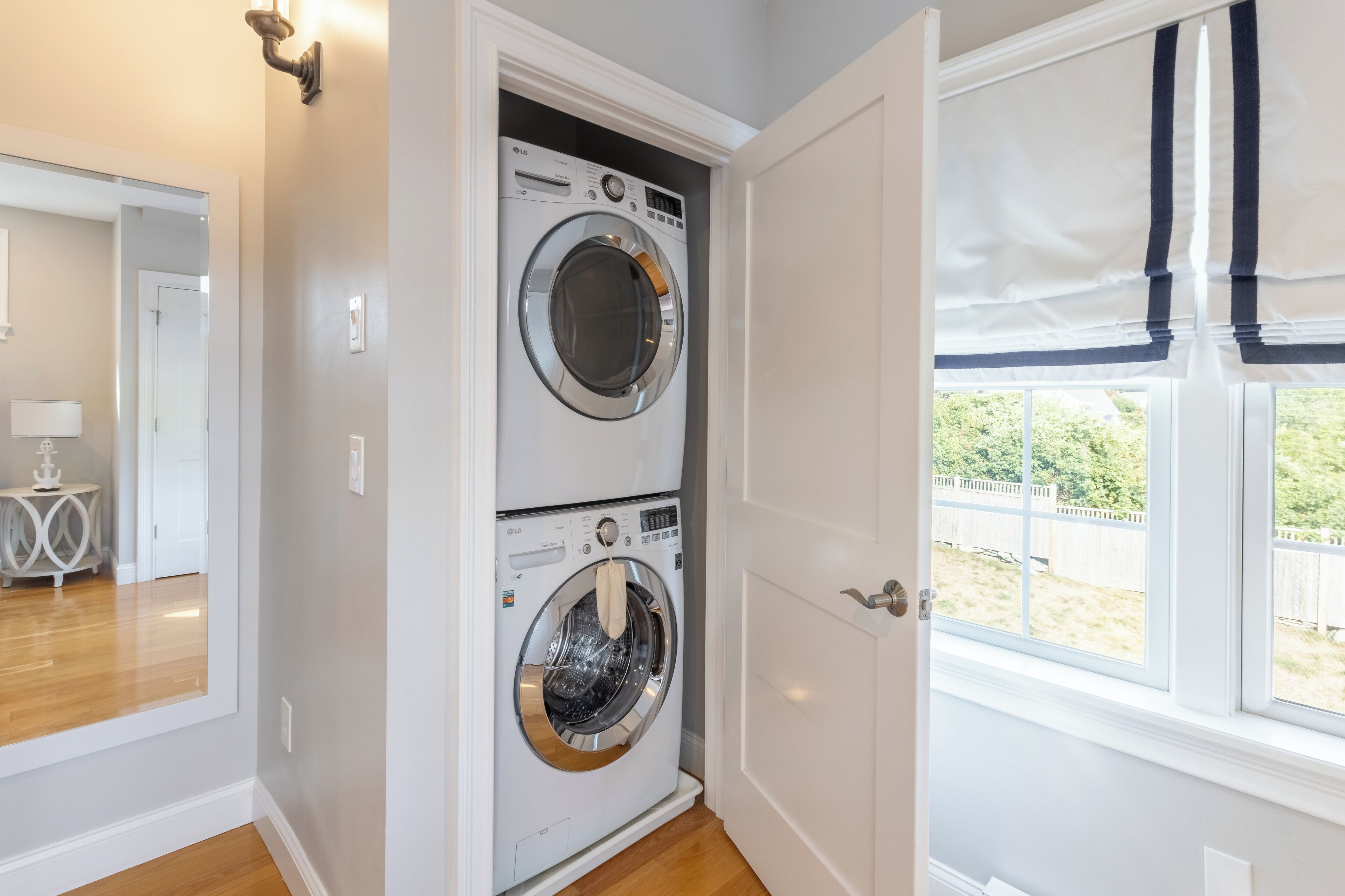 A laundry room with two stacked front-loading washing machines, a door leading to a hallway, and a window with a view of a fenced yard.