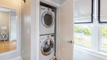 A laundry room with two stacked front-loading washing machines, a door leading to a hallway, and a window with a view of a fenced yard.