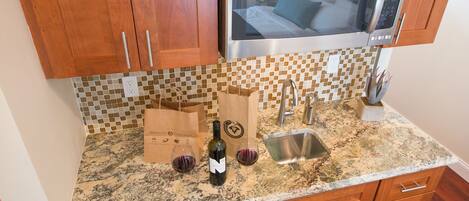A kitchen with wooden cabinets, a stainless steel microwave, a granite countertop, a sink, and wine glasses.