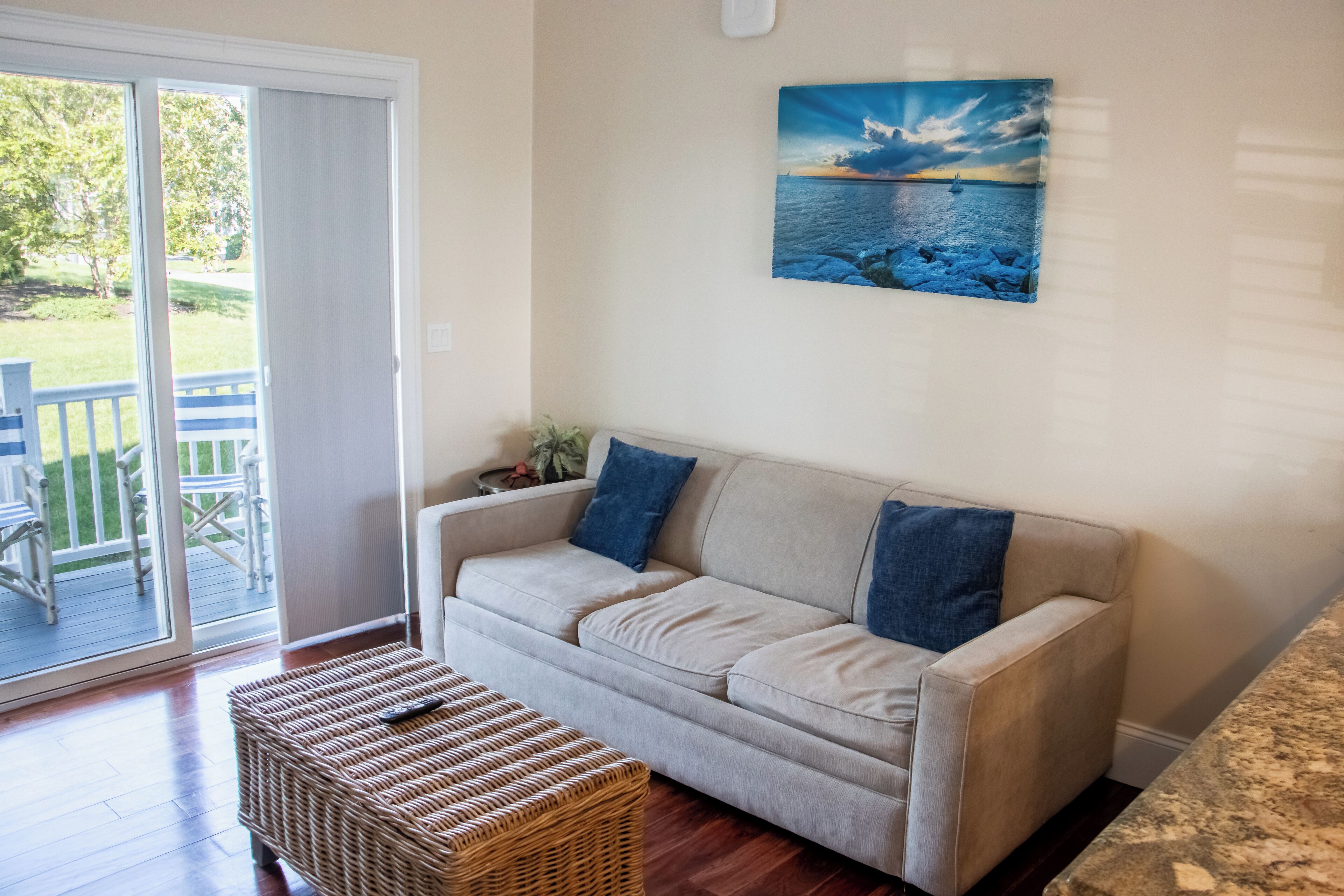 Un salon avec un canapé beige, un pouf en osier et un tableau représentant un paysage marin.