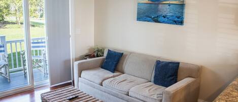 Un salon avec un canapé beige, un pouf en osier et un tableau représentant un paysage marin.