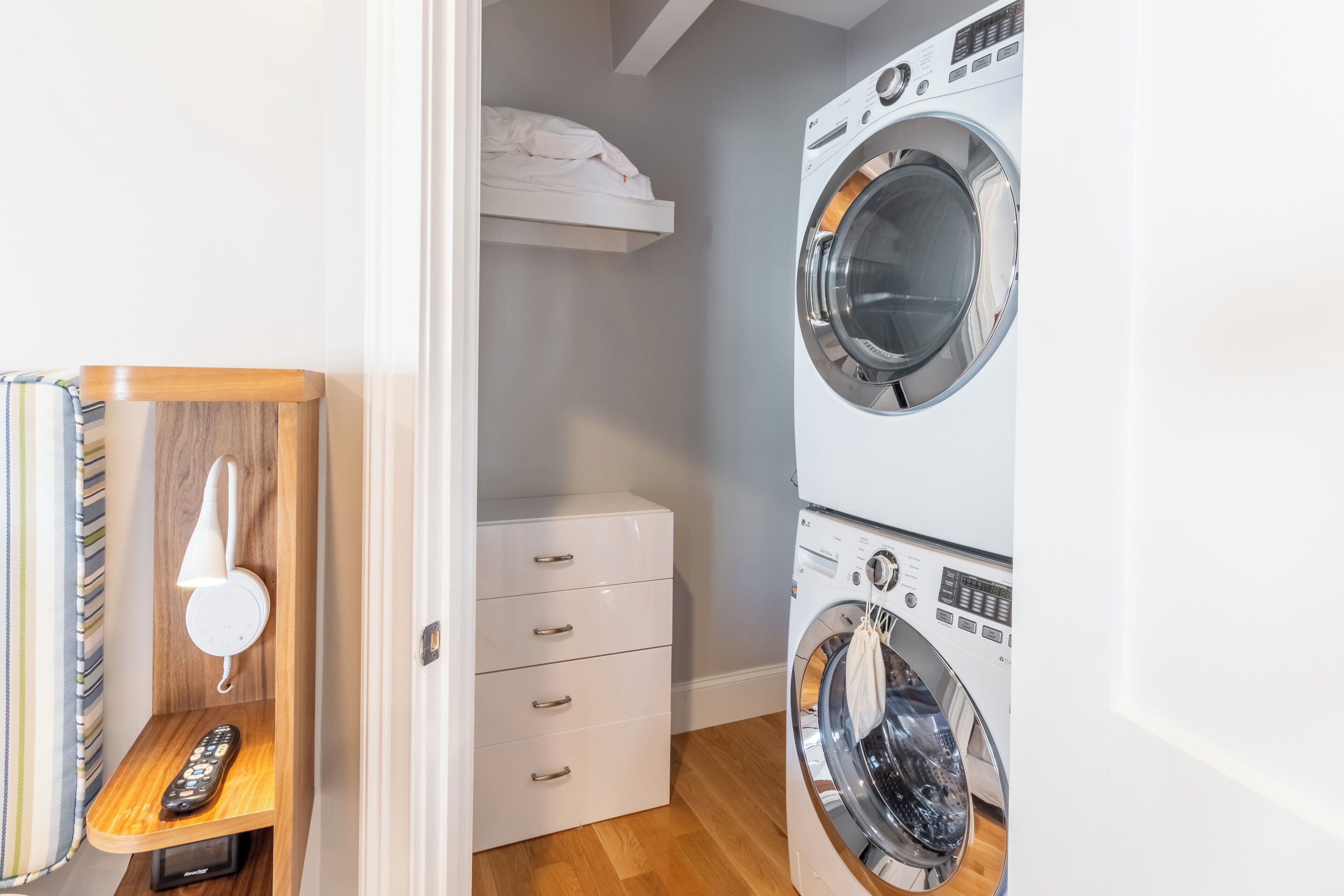 A laundry room with two stacked front-loading washing machines, a white cabinet with drawers, and a wooden shelf with a telephone and remote control.