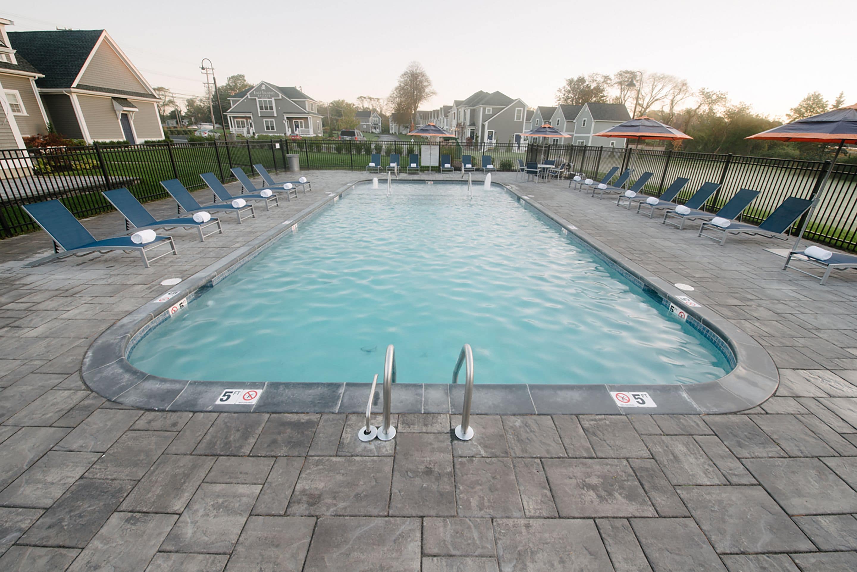 Ein rechteckiger Swimmingpool mit einer gefliesten Liegefläche, umgeben von Liegestühlen und Sonnenschirmen, und ein Zaun, der das Areal umschließt.