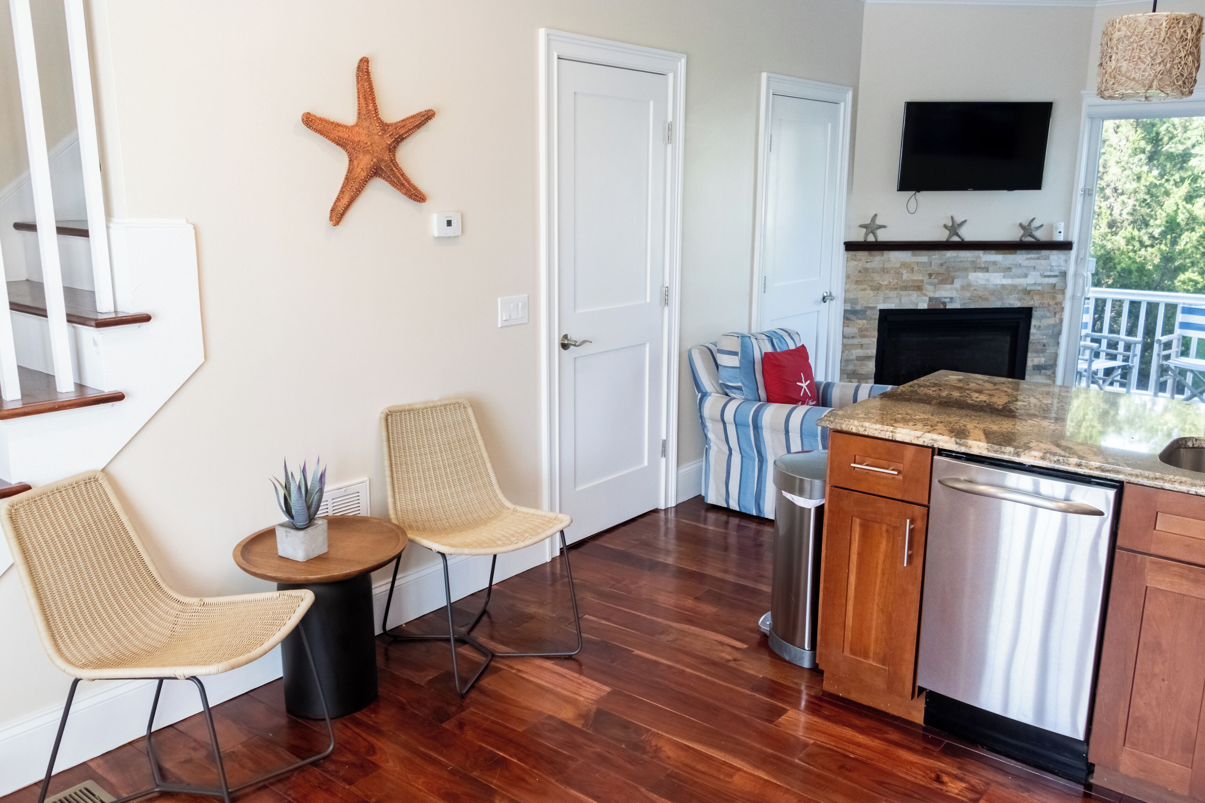 A kitchen with wooden flooring, a dining area with wicker chairs, a small round table, a built-in dishwasher, and a stone fireplace.