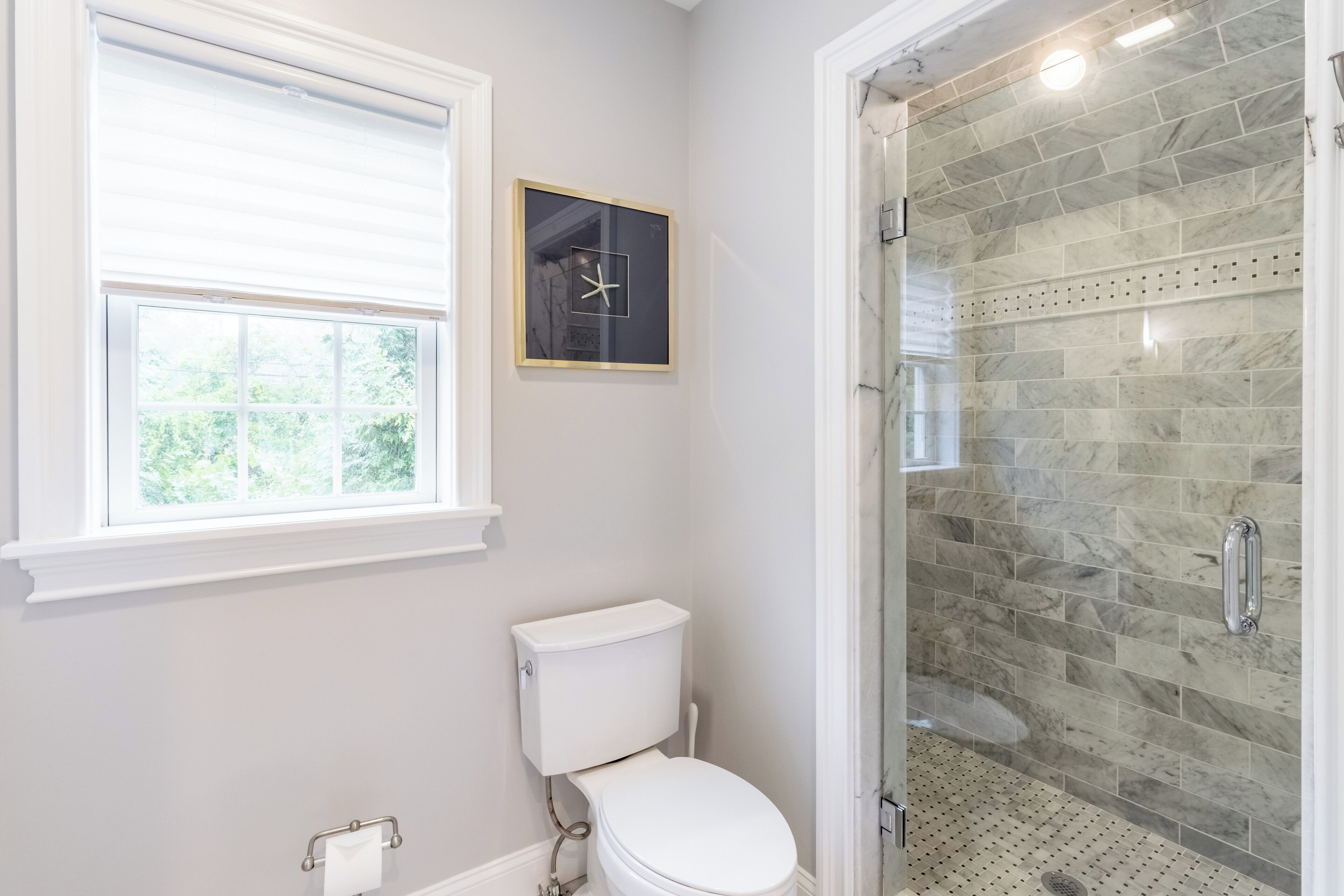 Un baño con inodoro, una ventana con persianas, un cuadro enmarcado y una ducha con puerta de cristal.