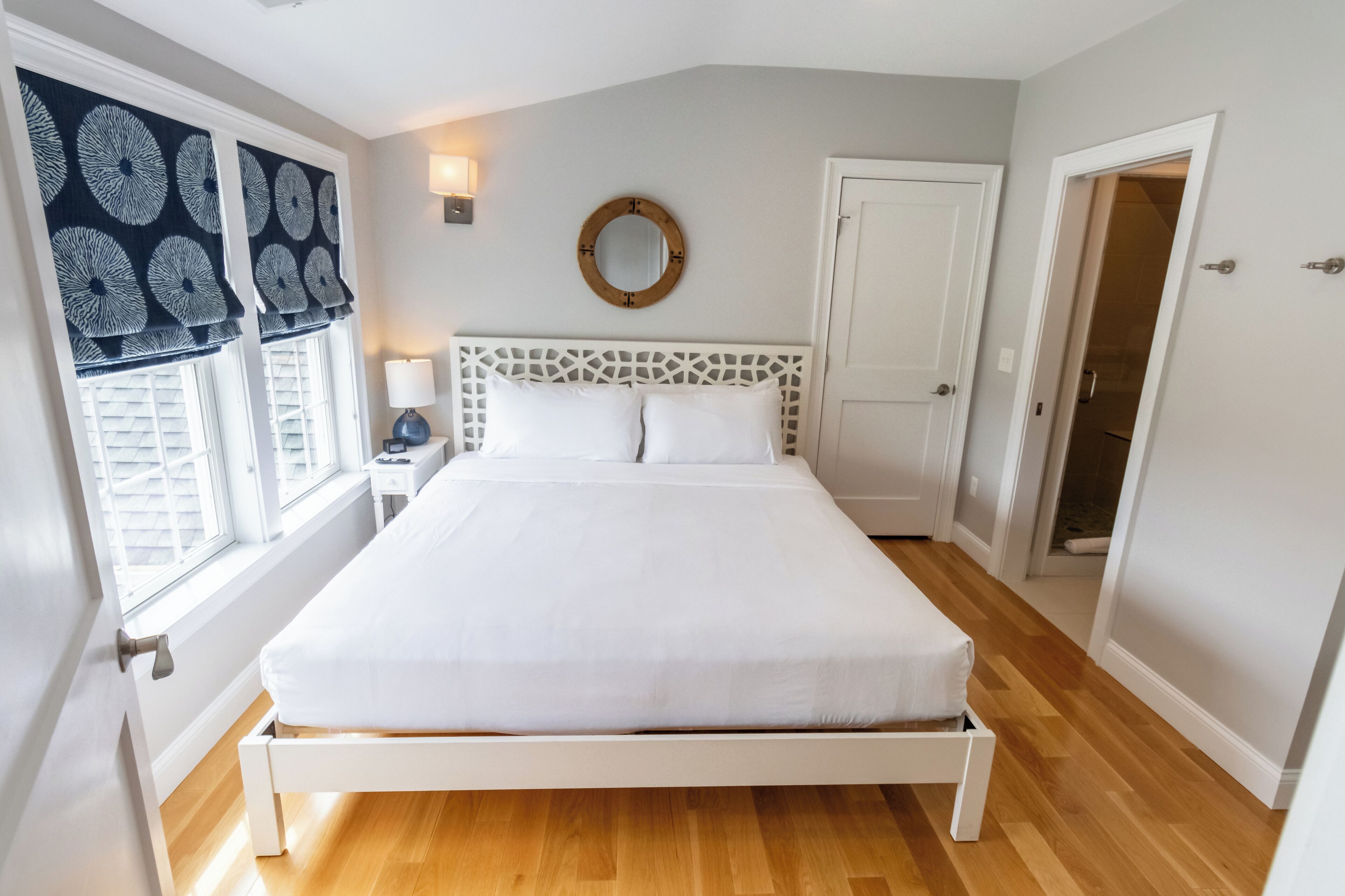 A bedroom with a white bed, a nightstand with a lamp, and a window with patterned curtains.