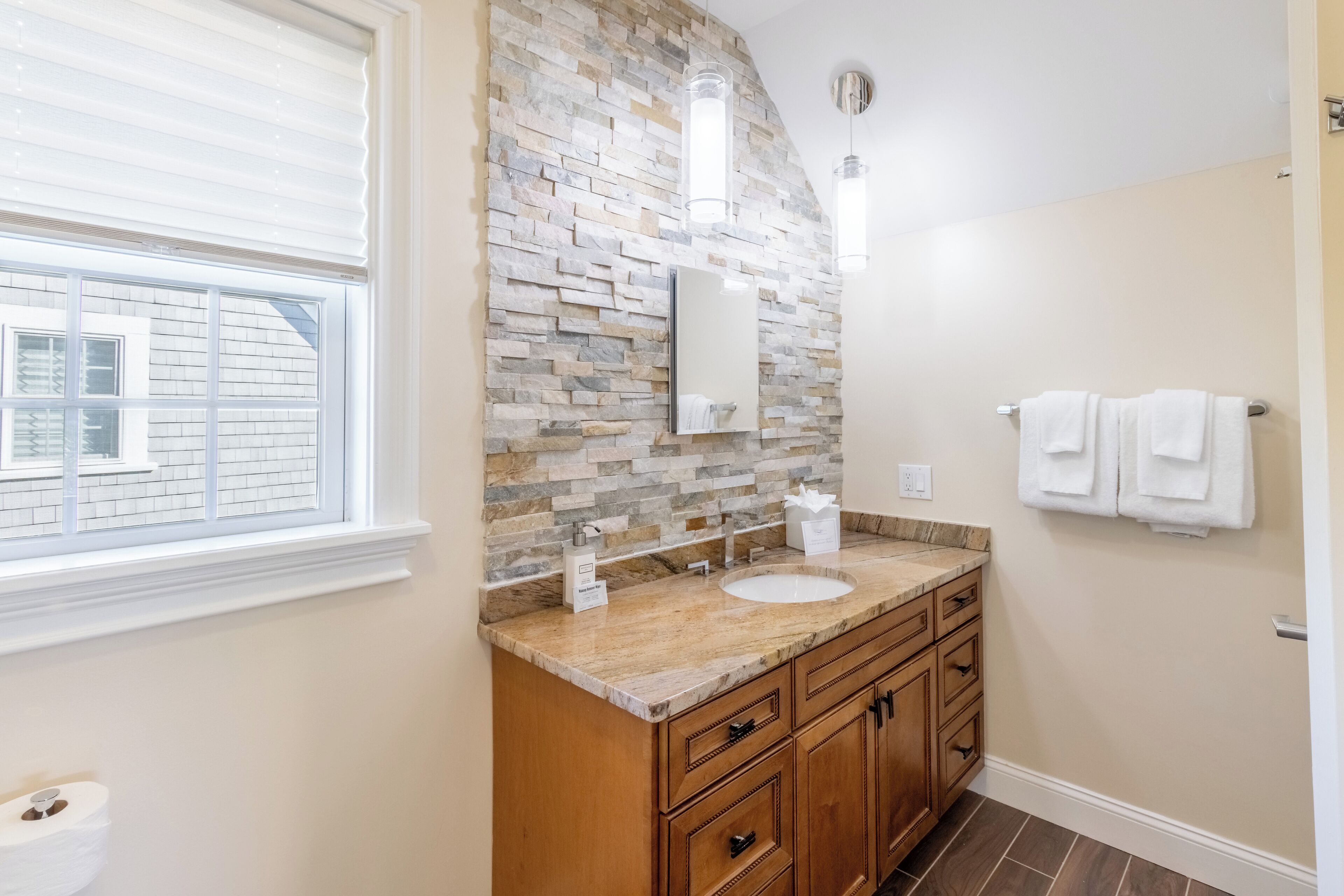 A bathroom with a stone backsplash, a sink, wooden cabinets, and a window with blinds.