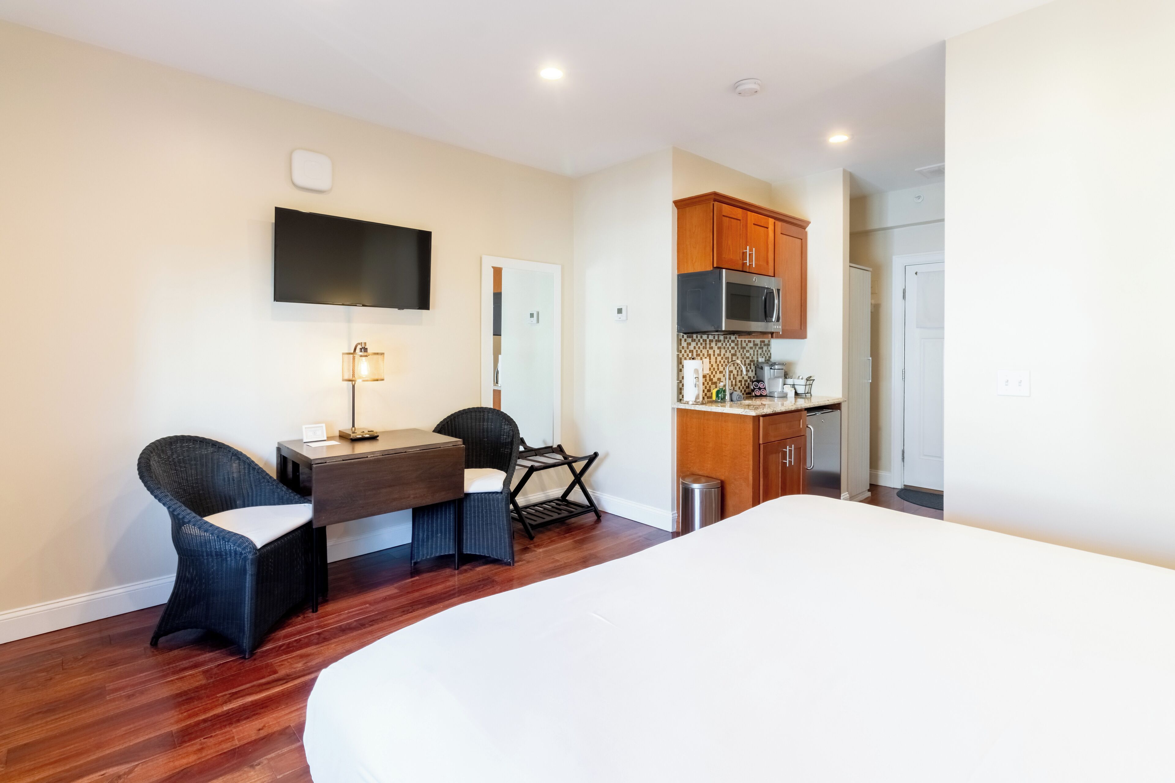 Habitación de hotel con cama, escritorio con dos sillas, televisor de pantalla plana en la pared, kitchenette con armarios de madera y microondas, y puerta que da a otra habitación.