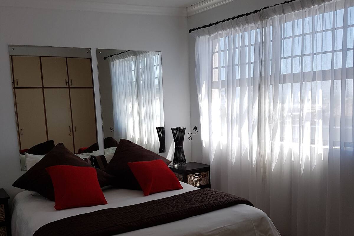 A neatly made bed with white and brown bedding, a bedside table with a vase, and a large window with sheer curtains.