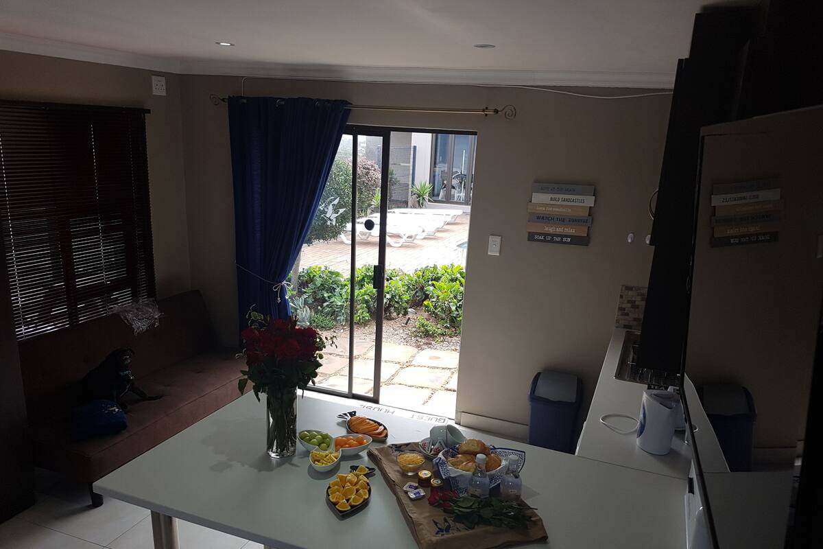 A kitchen with a dining table set with fruits and a vase of flowers. There is a window with a view of a garden and a blue curtain.