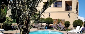 A pool area with lounge chairs, a small table, and a building with a beige facade and decorative elements. - Seagulls Guest House (Langebaan)