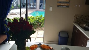 A dining area with a table set for breakfast, including fruits, pastries, and condiments. A vase with red flowers is on the table, and a view of a pool and outdoor seating is visible through the window.