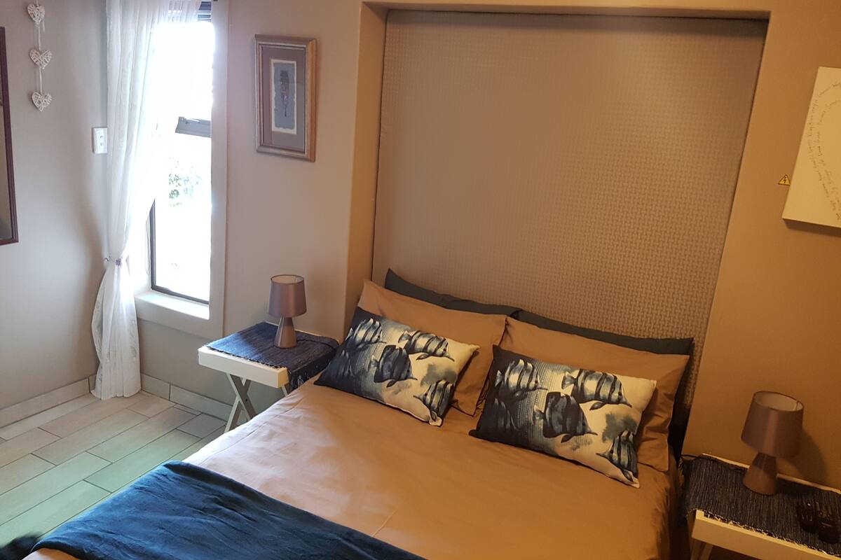 A bedroom with a bed, a nightstand, a lamp, a window with curtains, and a wall-mounted picture.