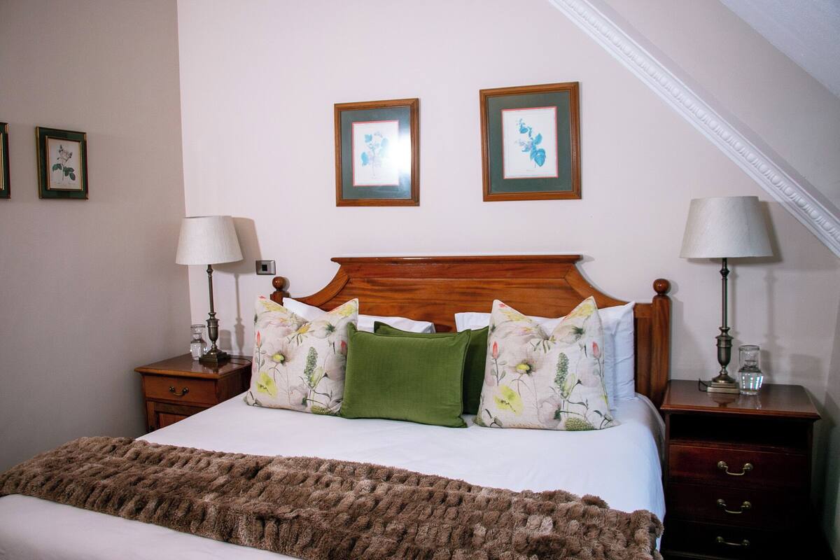 Cama bem arrumada com almofadas florais, cabeceira de madeira e duas mesas de cabeceira com abajures.