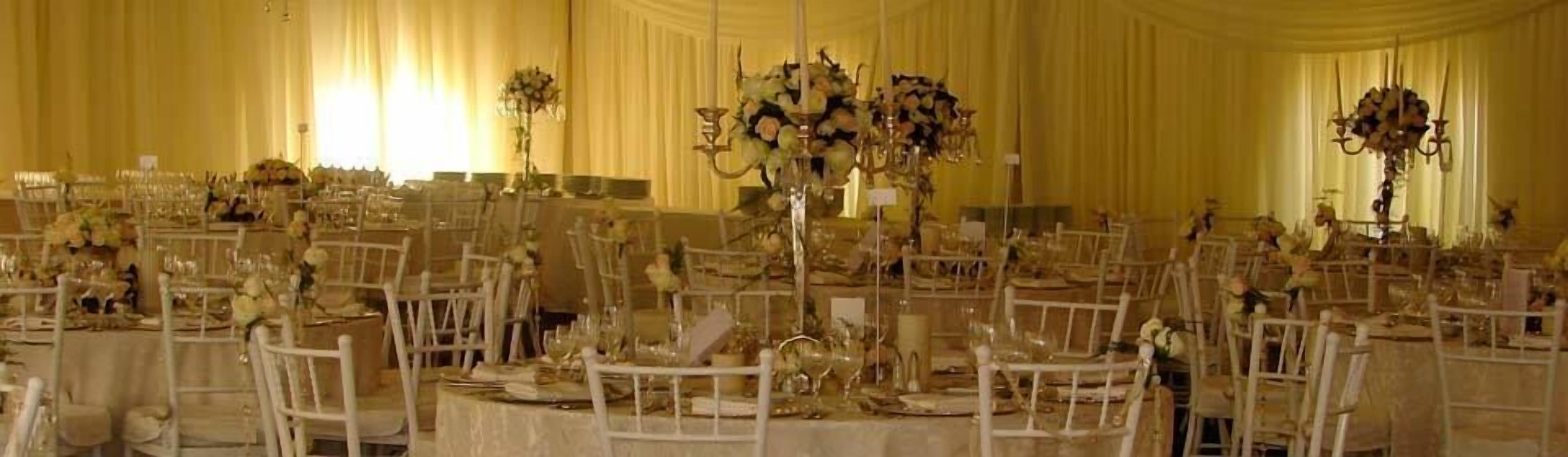 Un salón de banquetes con mesas redondas, sillas blancas y centros de mesa florales.