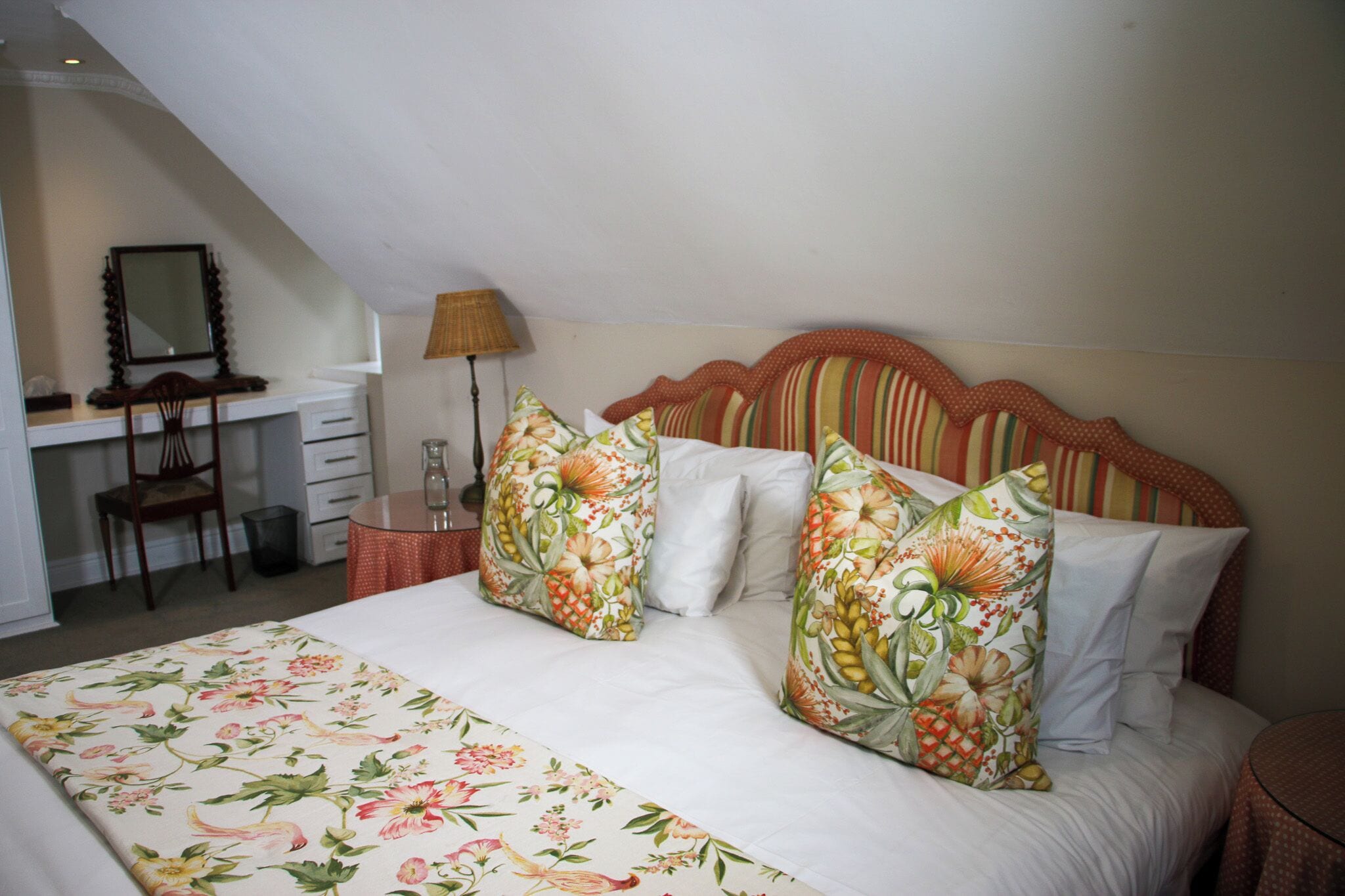 A bedroom with a bed, two pillows, a nightstand, a lamp, a chair, and a desk with drawers.