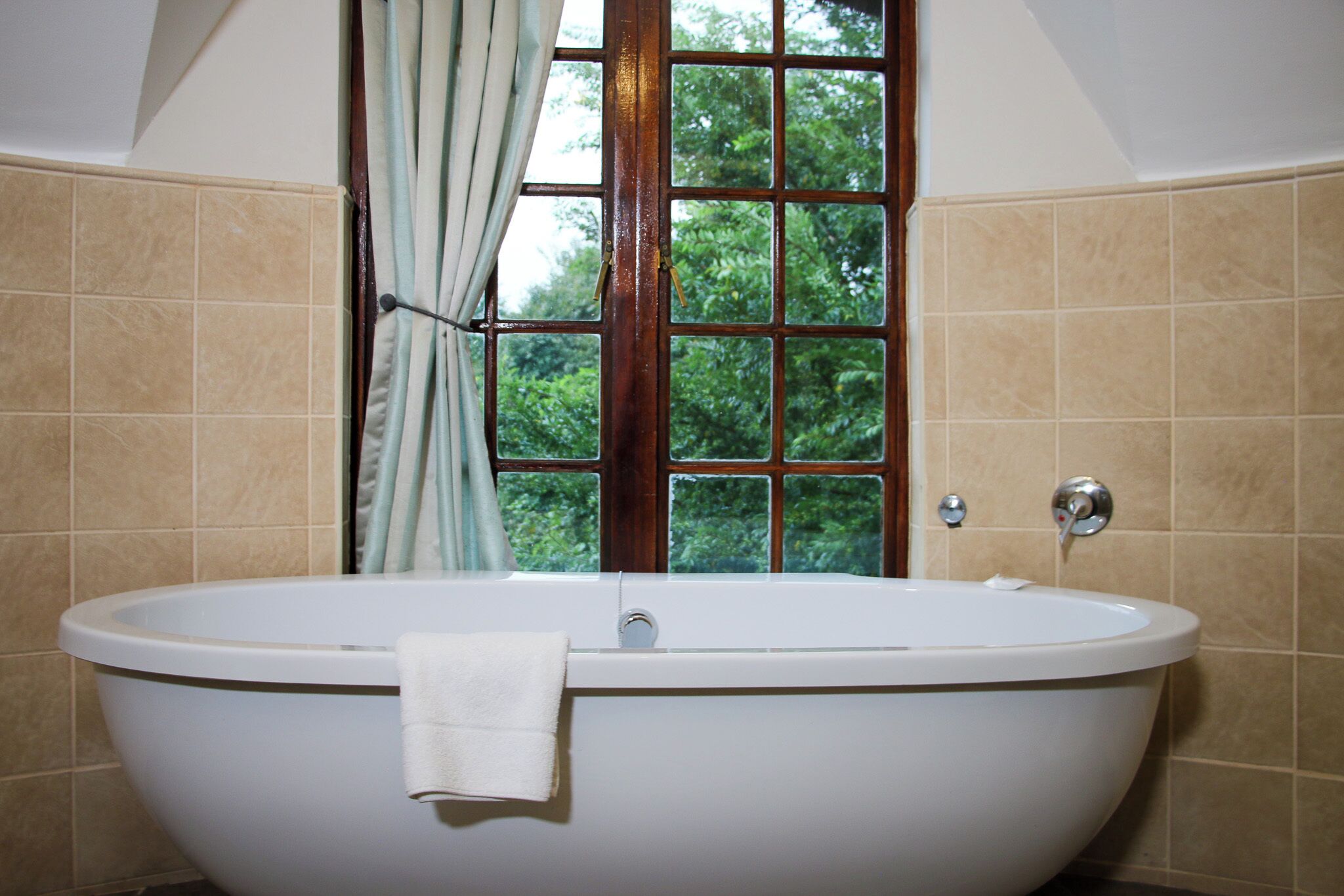 A bathroom with a freestanding bathtub, a window with curtains, and beige tiled walls.