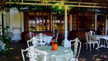 An outdoor seating area with white metal chairs and tables, surrounded by greenery and covered by a pergola.
