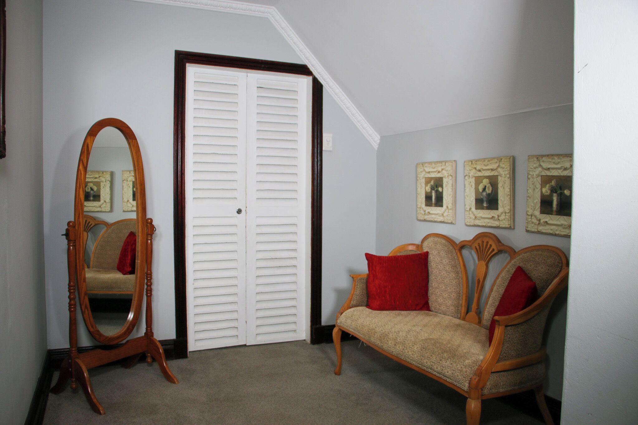 Una habitación con una silla de madera, un espejo, una puerta blanca y tres cuadros en la pared.